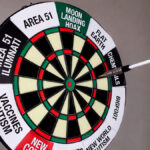 Dartboard labeled with conspiracy theories from AJ Gentile interview, including moon landing hoax and Area 51, with an arrow flying mid-air